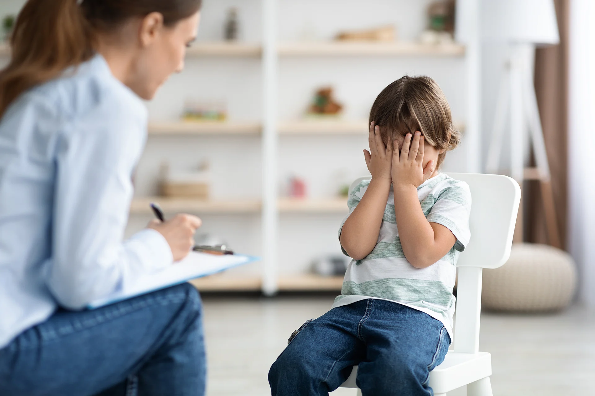 Psychological problems and help for kids. Upset little boy crying, covering face with hands, psychologist taking notes Psychologin hilft einem Kind. Das Kind verschänkt die Hände vor dem Gesicht.