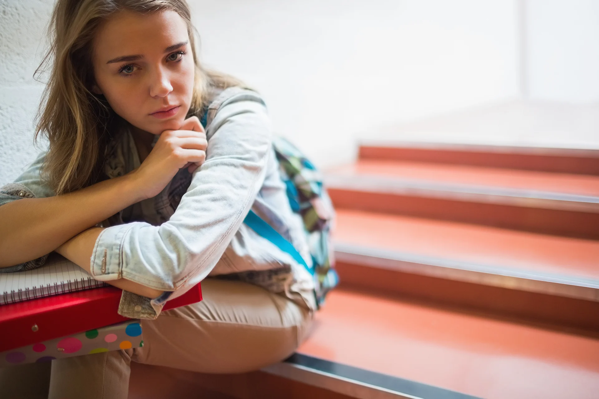 Eine Teenagerin sitzt traurig auf der Treppe in der Schule. Auf ihrem Schoß liegen Ordner und Hefter.